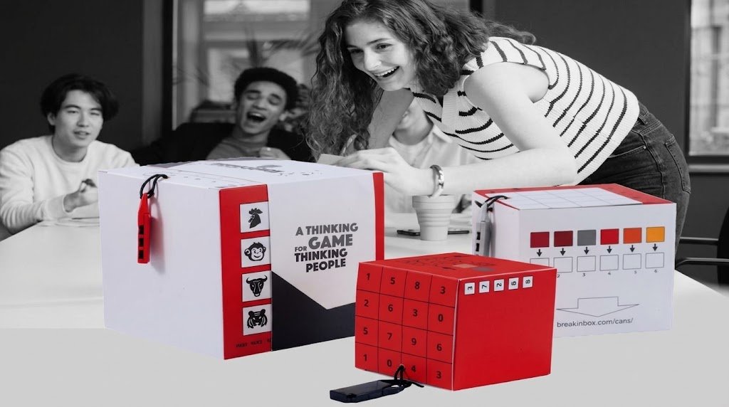 Three people laugh around a table as one of them opens a stack of red and white BreakInBox puzzle boxes